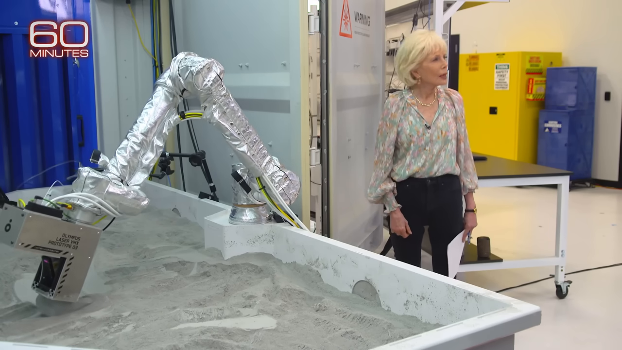 The Project Olympus robotic arm pointing a bright laser into a bed of grey simulated lunar dust.